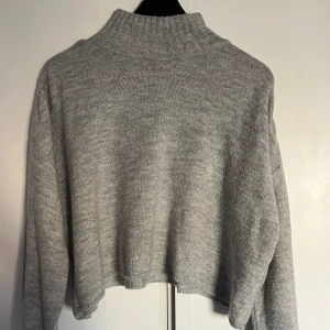 Grå stickad tröja från Gina Tricot - Säljer en mysig grå stickad tröja från Gina Tricot. Tröjan har en hög krage och är croppad med långa ärmar.