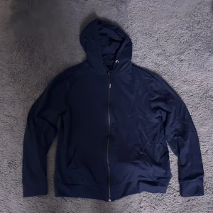 Mörkblå huvtröja med dragkedja från 157 - Säljer en mörkblå huvtröja från 157 med dragkedja framtill. Tröjan har en bekväm passform och är perfekt för kyligare dagar. Den har långa ärmar och en praktisk huva för extra värme. Perfekt för en avslappnad stil.