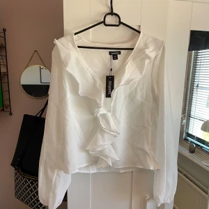 Vit blus  - Säljer en elegant vit blus från Boohoo med volangdetaljer framtill. Blusen har långa ärmar och en v-ringad design som ger en stilren look. Blusen är aldrig använd och har prislappen kvar! 