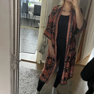 Mönstrad kimono  - Kimono med ett färgglatt paisleymönster i svart, orange och rosa. Kimonon har trekvartsärmar, fickor och ett knytband i midjan. Snygg både stängd och öppen med knytning baktill! Lent viskos satin tyg. Strl S/M. ord.pris 899:- 