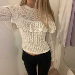 Topp med volangdetaljer - Säljer en vit blus med vackra volangdetaljer. Blusen har en tight passform och långa ärmar, perfekt för en stilren look. Den är tillverkad i ett fint hålstickat material som ger en romantisk känsla. Aldrig använd!