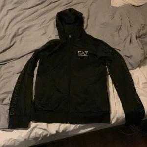 Svart tracktop från Emporio Armani - Säljer en stilren svart tracktop från Emporio Armani med dragkedja och EA7-logga på bröstet och ärmarna. Tröjan har långa ärmar och en bekväm huva, perfekt för en casual look.