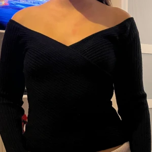 Svart offshoulder tröja - Snygg svart offshoulder tröja med omlott-design. Tröjan har långa ärmar och en ribbad struktur som ger en stilren look. Perfekt för en trendig outfit.