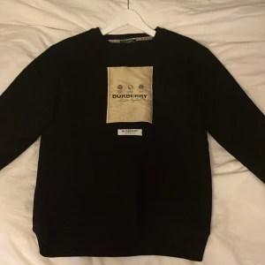 Svart tröja från Burberry - Snygg svart tröja från Burberry med en stor logga på framsidan. Tröjan har en klassisk passform och är perfekt för en stilren look. Den är långärmad och har ribbade muddar vid ärmslut och nederkant.