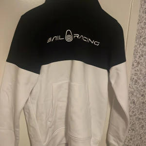 Svart och vit hoodie från Sail Racing - Snygg svart och vit hoodie från Sail Racing med logga på bröstet och ryggen. Hoodien har en klassisk passform med långa ärmar och en praktisk ficka framtill. Perfekt för en avslappnad stil. Modellen är Bowman och är köpt för omkring 1300-1400. Bara använd ett fåtal gånger och har inga som helst defekter. Skriv ifall du har några funderingar. 