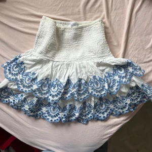 Vit kjol med blå broderier från Lindex - Charmig vit kjol från Lindex med vackra blå broderier längs volangerna. Kjolen har en elastisk midja för bekväm passform och är perfekt för en somrig look. Broderierna ger en extra touch av elegans och stil. Passar mig som har storlek xs!