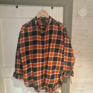 Rutig skjorta från Monki - Säljer en snygg rutig skjorta från Monki i svart, orange och vit. Skjortan har en avslappnad passform med långa ärmar och knappar framtill. Perfekt för en casual look!