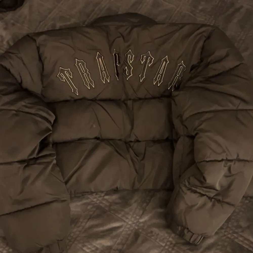 Snygg svart pufferjacka från Trapstar med dragkedja och logga på bröstet. Jackan har en cool design med text på ryggen och är perfekt för kyligare dagar. Den är långärmad och har en modern look. Pris kan diskuteras vid snabbt köp. Takit.