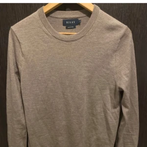 Beige tröja från Riley - Säljer en stilren beige tröja från Riley i merinoull. Tröjan har en rund halsringning och långa ärmar, perfekt för kyligare dagar. Den är mjuk och bekväm, vilket gör den till ett utmärkt val för både vardag och mer uppklädda tillfällen.