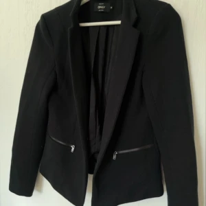 Svart kavaj från Only - Snygg svart kavaj från Only med dragkedjedetaljer på fickorna. Perfekt för en stilren look. Kavajen har långa ärmar och en klassisk krage.