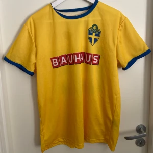 Sverige fotbollströja  - Fotbollströja från svenska landslaget i storlek S/M med blå detaljer vid hals och ärmslut. Tryckt Bauhaus-logga på bröstet och ryggen samt nummer 22 i blått. Materialet är lätt och andas. Klassisk landslagsdesign med broderat Sverigemärke.