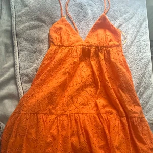 Orange spetsklänning från Zara - Superfin orange kortklänning från Zara med broderade blommor i spets, smala axelband och v-ringning. Klänningen har öppen rygg med knyt och volang nedtill. Perfekt för sommaren! 