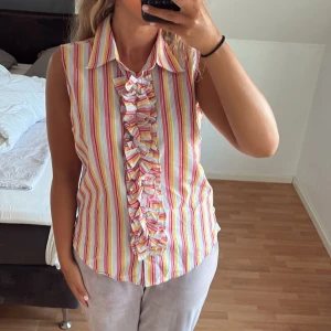 Randig ärmlös skjorta med volang - Säljer en färgglad ärmlös skjorta med vertikala ränder i rosa, gul, vit och orange. Skjortan har klassisk krage och snygga volangdetaljer längs knappraden framtill. Perfekt för dig som gillar en lekfull och somrig stil. Aldrig använd då den är för stor!