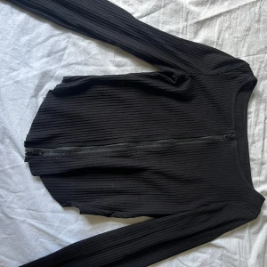 Svart ribbad kofta med dragkedja - Svart ribbad kofta med lång ärm och dragkedja framtill. Tröjan har en tight passform och är tillverkad i ett stretchigt, ribbat material som sitter snyggt på kroppen. Perfekt att slänga över en topp eller bära stängd för en clean look.