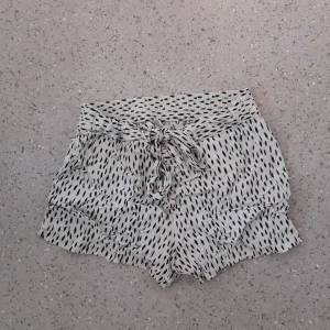 Mönstrade shorts från Jacqueline de Yong - Snygga vita shorts med svart mönster från Jacqueline de Yong. De har hög midja, bred resår och knytband framtill. Lätta volanger längs benen ger en chill vibe. Perfekta för varma dagar när du vill vara både bekväm och trendig. OBS! Liten i storleken