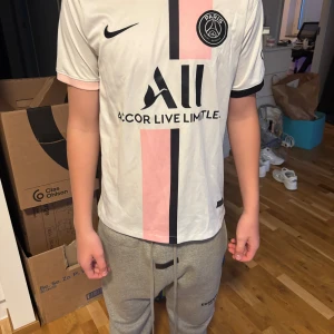 PSG vit och rosa fotbollströja Nike M - Modell är 184, och väger 68. Snygg PSG fotbollströja från Nike i vitt med en rosa och svart vertikal rand på framsidan. Tröjan har korta ärmar med svarta och rosa muddar, broderad Nike-logga och PSG-märke på bröstet. Tillverkad i lätt och ventilerande Dri-FIT material.