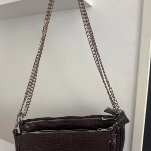 Brun axelväska med kedja och croc-mönster - Snygg mörkbrun axelväska i croco-imitation med silverfärgad kedjerem. Väskan har två stora fack med dragkedja och ett mindre innerfack. Väskan är i använt skick, men går fortfarande att användas och kan fräschas till för att se ännu bättre ut.