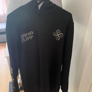 Svart hoodie Sana Duri Hardstyle - Svart hoodie från Sana Duri med tryck på bröstet och ärmen samt stor grön och grå dödskallegrafik och texten 'HARDSTYLE BABY!' på ryggen. Klassisk känguruficka och huva, perfekt för dig som gillar streetwear och festivalvibe.