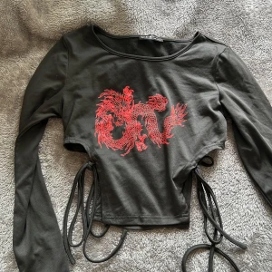 Svart croppad topp med röd drake - Cool svart croppad topp från SHEIN med lång ärm och snörning i sidorna. Framsidan har ett stort rött drakmotiv som verkligen sticker ut. Toppen är figurnära och gjord i mjuk bomullsmix, perfekt för dig som vill ha en edgy stil.