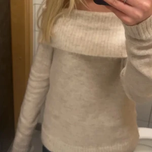 Beige offshoulder stickad tröja - Supermysig beige stickad tröja med bred ribbad kant och offshoulder-modell. Perfekt för dig som gillar en chill men ändå snygg stil. Långärmad och i mjukt material som håller dig varm under kyliga dagar.