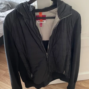 Svart quiltad jacka med huva - Svart jacka/cardigan från pjs med quiltade partier framtill och på ryggen, samt mjuka ärmar. Jackan har huva, dragkedja och två fickor med dragkedja. Snygg patch på ena ärmen ger extra attityd. Perfekt för dig som gillar en sportig och urban stil. Den har ett pyttelitet hål på magen som ej syns!