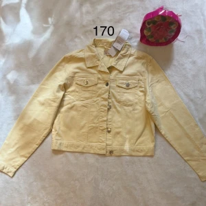 Barn Ljusgul jeansjacka från UO - Fräsch ljusgul jeansjacka från UO med klassisk krage, silverfärgade knappar och två bröstfickor med lock. Jackan har en rak passform och är tillverkad i bomull, perfekt för vår och sommar. Snygg att styla över en t-shirt eller klänning.