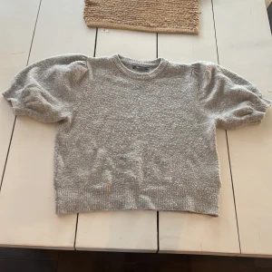 Grå stickad tröja från Gina Tricot - Supermjuk grå stickad tröja från Gina Tricot i storlek XS. Tröjan har korta puffärmar och ribbade muddar vid hals, ärmslut och nederkant.