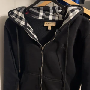 Svart Burberry hoodie  - Svart hoodie från Burberry med klassisk rutig insida i huvan och längs dragkedjan. Tröjan har huva med snörning, hel dragkedja och långa ärmar. Ikonisk Burberry-logga broderad på bröstet. Perfekt för dig som gillar stilrena och lyxiga streetwear-plagg. Pris kan diskuteras! Passar XS också! Nyskick!