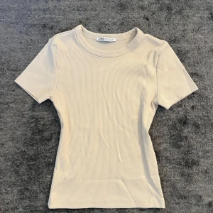Beige ribbad topp från Zara - En stilren beige ribbad topp från Zara med korta ärmar och rund halsringning. Toppen har en slim passform och är tillverkad i mjuk bomull, perfekt för en clean och enkel look. Passar till allt och är superenkel att styla.