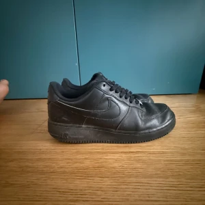 Nike Air Force 1 helsvarta sneakers - Klassiska Nike Air Force 1 i helsvart läder med svart sula och svarta skosnören. Snygg och tidlös modell med låg profil, rund tå och diskret Nike-logga på sidan. Perfekta för dig som gillar en clean och stilren look.