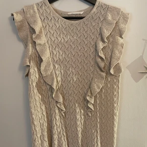Beige stickad blus med volang - Supersöt beige stickad blus med volangdetaljer längs axlarna och ärmlös design. Guldglittrig! Storlek 164, skulle säga S!