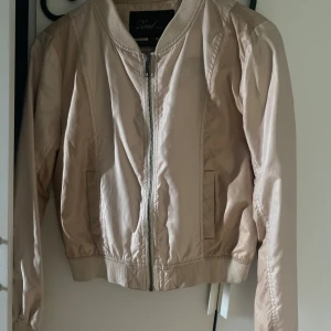 Beige bomberjacka från Zoul Edition - Snygg beige bomberjacka från Zoul Edition i storlek 38. Jackan har klassisk bombermodell med ribbade muddar, dragkedja framtill och två fickor. Materialet är glansigt och lätt, perfekt för vår och höst. Enkel att matcha till olika outfits.