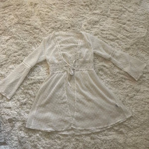 Vit ”kofta” med spets från SHEIN - Supersöt vit ”kofta” från SHEIN med prickigt mönster, spetsdetaljer i midjan och vid halsringningen. Klänningen har långa utsvängda ärmar och knytning framtill. Perfekt till stranden över bikinin!🩷