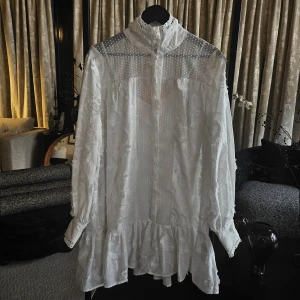 Custommade klänning - Elorie - Superfin vit kortklänning med broderade blommor, spetsdetaljer och hög krage. Klänningen har långa ärmar, knappar framtill och volang nertill. Perfekt för dig som gillar romantisk och vintage-inspirerad stil.