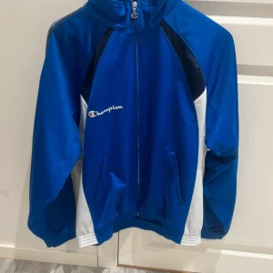 Blå Champion jacka med logga - Snygg blå vindjacka från Champion med vita och marinblå detaljer. Jackan har dragkedja med coolt C-drag, broderad Champion-logga på bröstet och tryck på ryggen med 'Polisportiva Virgilio Volley'. Perfekt för dig som gillar sportig stil.