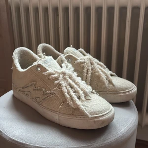 Beige Zadig Voltaire skor med fransiga detaljer - Snygga beige sneakers från Zadig & Voltaire i grov bomull med fransiga snören och råa kanter. Skorna har en vit platt sula och en cool blixtdetalj på sidan. Perfekta för dig som gillar en avslappnad och trendig stil. PRIS GÅR ATT DISKUTERA 
