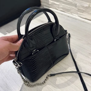 Svart handväska med krokodilmönster - Snygg svart handväska med krokodilmönster och silverfärgad kedjedetalj. Väskan har både handtag och en avtagbar axelrem, samt en dragkedja för säker förvaring. Perfekt för att ge en stilren touch till din outfit. Zara