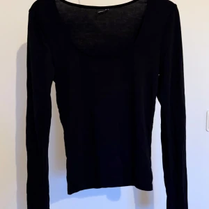 Svart långärmad topp med rund hals - En svart långärmad topp med rundad, djup urringning, med slits vid ärmslutet. Tunt och mjukt material som känns skönt mot huden. Enkel och stilren design som funkar året runt. 