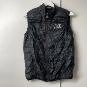 Svart dunväst från EA7 Emporio Armani - Svart quiltad dunväst från EA7 Emporio Armani med hög krage och dragkedja framtill. Västen har en stilren design med logga i silver på bröstet och två fickor. Perfekt för lager-på-lager och snygg till streetwear. Finns ett jätte litet hål på sidan därför jag säljer den billigt 