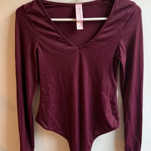 Vinröd ribbad topp från Hunkemöller - Säljer en vinröd långärmad topp/body från Hunkemöller med v-ringning och ribbad struktur. Toppen har en figurnära passform och är tillverkad i ett mjukt, stretchigt material som känns skönt mot huden. Perfekt för dig som gillar stilrena och bekväma plagg.