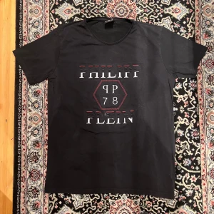 Svart Philipp Plein t-shirt med tryck - Svart t-shirt från Philipp Plein i bomull med stort vitt och rött tryck på bröstet. Snyggt grafiskt mönster med texten 'PHILIPP PLEIN' och PP78 i en sexkant. Klassisk passform och rund hals. Perfekt för dig som gillar streetwear med lyxig vibe. Väldigt snygg har en lätt fläck som går bortom tvätten