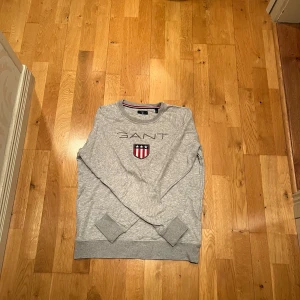 Grå sweatshirt från Gant. - Snygg grå sweatshirt från Gant med broderad logo och sköld på bröstet. Tröjan har rund halsringning med röd och marinblå rand på insidan, samt ribbade muddar vid ärmslut och nederkant. Perfekt för en chill och stilren look.