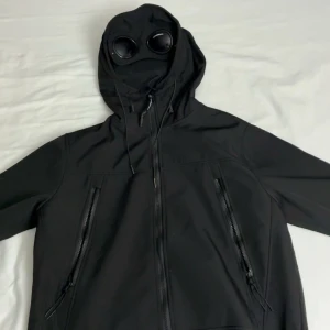 Svart Jacka från C.P. Company - Svart softshelljacka från C.P. Company med deras ikoniska goggles i huvan – en riktig statement-piece för dig som gillar en unik och stilsäker streetwear-look. Jackan har dragkedjestängning framtill, två snedställda fickor och en justerbar huva med snörning för perfekt passform. Endast använd vid två tillfällen – i mycket fint skick! Storlek M, men passar även dig som vanligtvis bär S. Först till kvarn! 