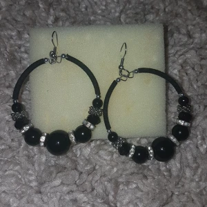 Stora svarta örhängen med pärlor - Snygga stora örhängen i svart med runda pärlor och silvriga detaljer. De har en cirkelformad design och glittrande inslag mellan pärlorna som ger extra bling. Perfekta för dig som vill sticka ut med ett coolt statement-smycke. PRIS KAN DISKUTERAS!