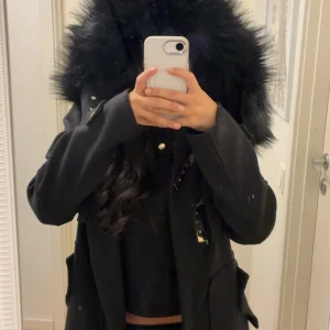 Svart parkajacka med fuskpäls från DM - Svart parkajacka från Dennis Maglic med stor huva kantad av fluffig svart fuskpäls. Jackan har snygga gulddetaljer, dragkedja framtill och dekorativa spännen. Perfekt inför höst/vinter. Köpt för 1799kr och säljs inte längre i hemsidan. 
