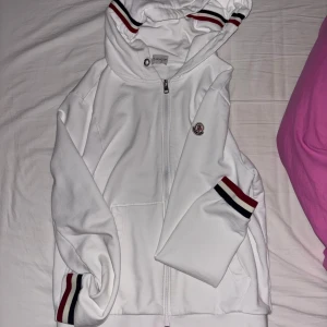 Vit Moncler hoodie med dragkedja - Vit hoodie från Moncler med dragkedja framtill och huva. Ribbade muddar och huva med röd, blå och vit randig detalj. Bild på QR-kod kan skickas vid intresse, snörena från hoodien ingår, kan skickas bild på dom också , storlek XL nypris ca 5000kr