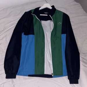 Lacoste Sport vindjacka XS - Snygg vindjacka från Lacoste Sport i färgblock med marinblått, grönt och blått. Jackan har dragkedja framtill, elastiska muddar och stor Lacoste-logga på ryggen. Perfekt för dig som gillar sportig och clean stil.