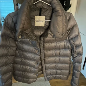 Grå Moncler dunjacka med hög krage - Säljer en snygg grå dunjacka från Moncler med hög krage och quiltad design. Jackan har en stilren look med blanka tryckknappar och dragkedja. Perfekt för kalla dagar när du vill hålla stilen och värmen. Passar dig som gillar exklusiva märken och klassisk vibe.