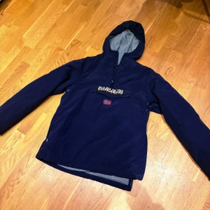 Mörkblå vindjacka från Napapijri - Säljer en mörkblå vindjacka från Napapijri med huva och halv dragkedja. Jackan har en stor ficka framtill med logga och norsk flagga. Perfekt för blåsiga dagar och snygg streetstil. Fodrad insida och långärmad modell.