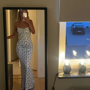 Vit långklänning med blå blommor - Supersöt långklänning i vitt med blått blommönster och smala axelband som knyts på axlarna. Klänningen har en figurnära passform och snygg snörning framtill vid bysten. Aldrig använd